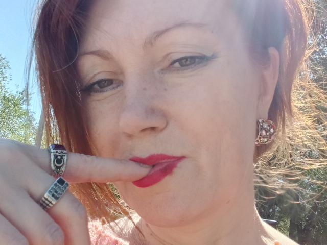 SadieLorri with red lipstick, brown hair, and green eyes, touching her finger to her red lips, wearing a silver ring and jeweled earrings, sunlit face, slight smile.