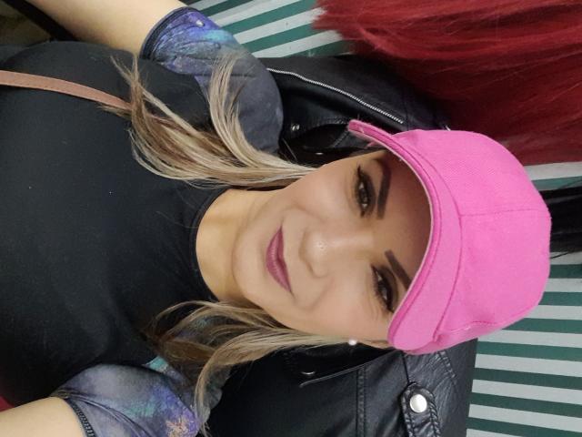 ChantalHotX met blond haar, een roze pet, een zwart shirt en glanzende paarse lippenstift, draagt een glimmende zwarte jas, toont een schattige glimlach, met zichtbare wenkbrauwen en subtiele make-up..