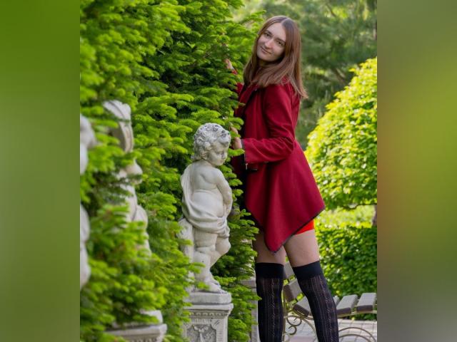 TeniOfLove with brown hair, red dress, and black thigh-high socks, leaning on a white baby statue, greenery in background.
