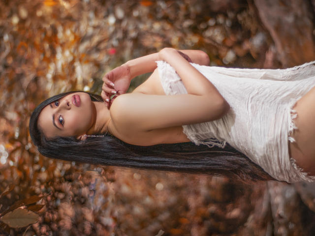 HannaRobertss with long black hair, wearing a white, lacy, strapless dress, looking over her shoulder, showing her ass, in a forest with autumn leaves.