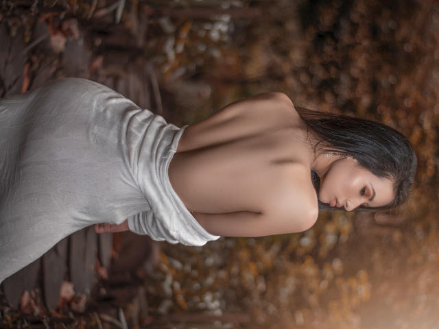 HannaRobertss with bare back, long black hair, and white dress, standing with ass towards camera, looking down, in forest. Her tits are not visible. Her pussy is not visible.