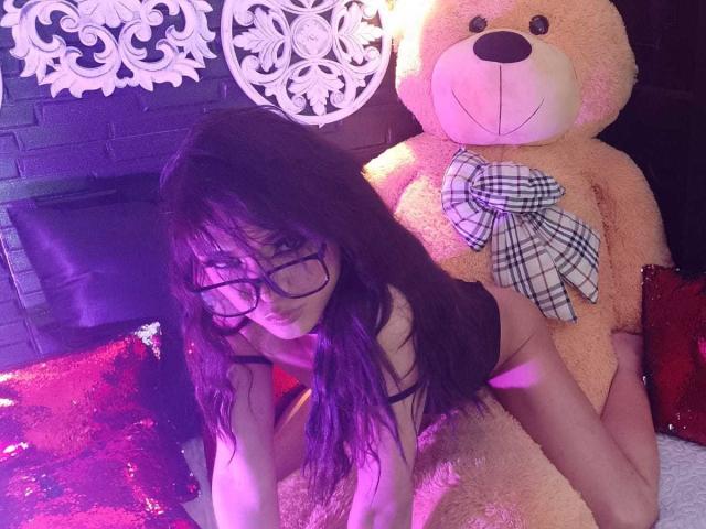 ChantalSophie mit langem, dunklem Haar, Brille und schwarzer Spitze, liegt auf einem Bett mit einem riesigen Teddybären, einer karierten Schleife, einem roten, mit Pailletten besetzten Kissen und violetter Beleuchtung. Brüste sind sichtbar, das Gesäß ist leicht angehoben..