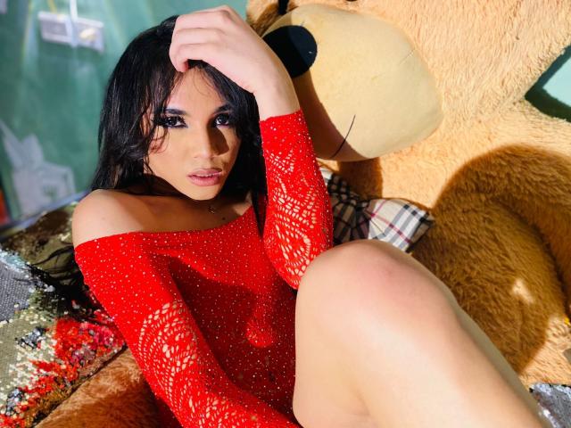 ChantalSophie, con cabello negro largo, vestida con una blusa roja brillante con un hombro descubierto, sentada contra un enorme oso de peluche, con la mano en el pelo, mostrando sus pechos y glúteos..