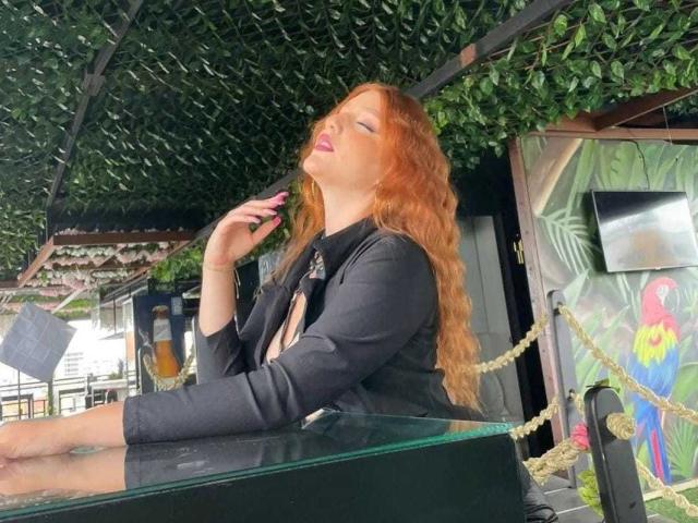 MelisaLee, con cabello rojo, chaqueta negra y lápiz labial morado, sentada, con los ojos cerrados, tocándose el cuello, una mesa de vidrio frente a ella, un techo verde y frondoso arriba. Sus senos apenas son visibles..