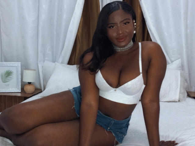EmmaBlosson with dark skin, wearing a white lace bra, showing her big boobs, and denim shorts, sitting on a bed, looking at the camera, with a choker necklace.