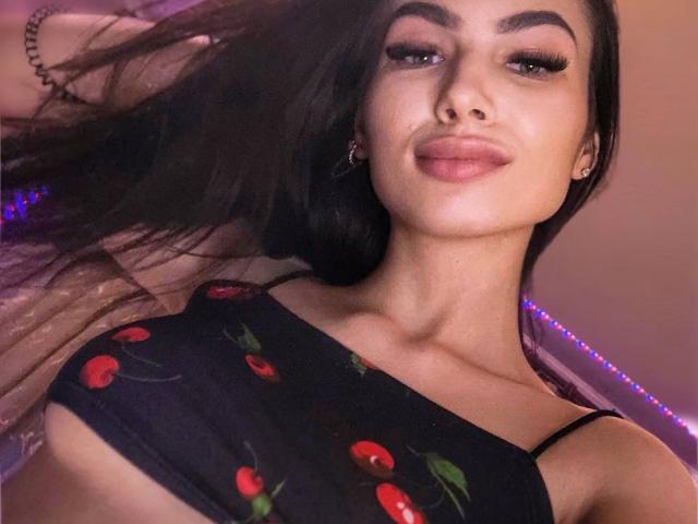 FluffyQBunny, avec ses cheveux foncés, ses lèvres pulpeuses et ses longs cils, porte un crop top noir avec des motifs de cerises, laissant entrevoir ses seins. Elle regarde l'appareil photo avec un léger sourire..