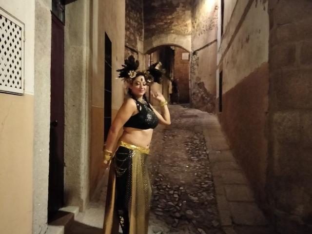 WetSweetKim con reggiseno nero, catene dorate, gonna nera e dorata e un copricapo piumato, in piedi in un vicolo acciottolato di notte, che mostra il seno e i glutei..