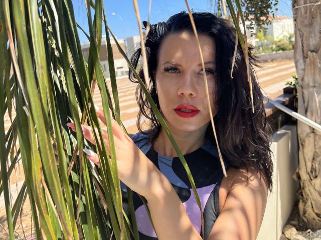 JulietteDolce, com lábios vermelhos, olhos azuis e cabelo escuro ondulado, em pé em frente a folhas verdes altas, vestindo uma blusa preta e roxa com estampas, iluminada pelo sol, ao ar livre..