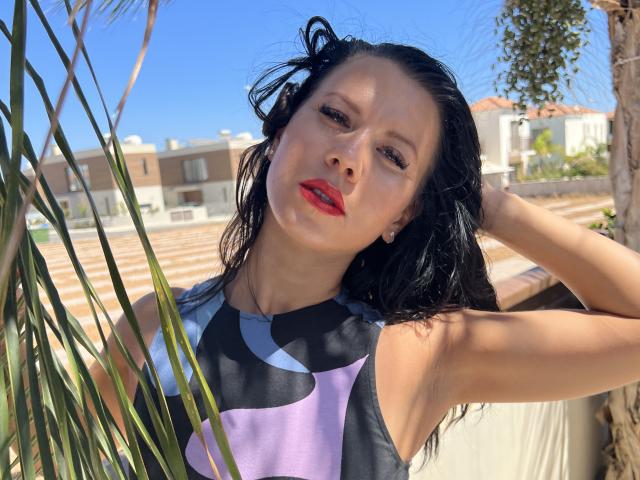 Juliette Dolce mit nassen, schwarzen Haaren, rotem Lippenstift und einem schwarzen und violetten Kleid ohne Ärmel, steht neben Palmenblättern, blickt nach oben und hat eine Hand in ihrem Haar..