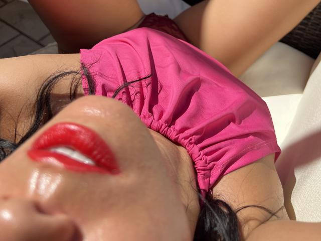 JulietteDolces Gesicht mit glänzenden roten Lippen, schwarzem Haar und einem rosafarbenen Oberteil, liegend, im Sonnenlicht, wobei ihre Brust und Brustwarzen zu sehen sind, trägt sie rote Spitzenunterwäsche, in einer entspannten, sonnenbeschienenen Position..