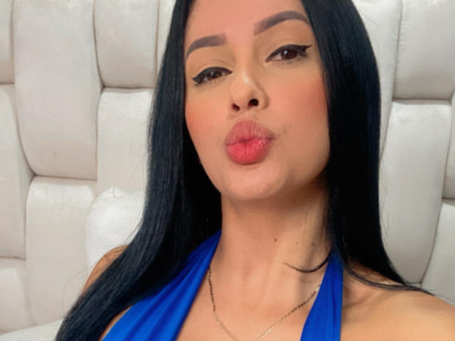 EmilyParkerr with long black hair, wearing a blue halter top, pouting her pink lips, brown eyes, and wearing a thin necklace, against a white, tufted background.