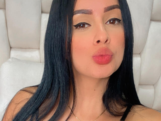 EmilyParkerr has black hair, brown eyes, and red lipstick, pouting her plump ass. She's wearing a black top and a gold necklace, sitting on a white cushion.