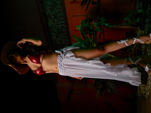 HannaRobertss stands in dim light, wearing a red metal bra, white frilled skirt, and brown hat, with her hand on her hat, showing her tan skin and bare ass.