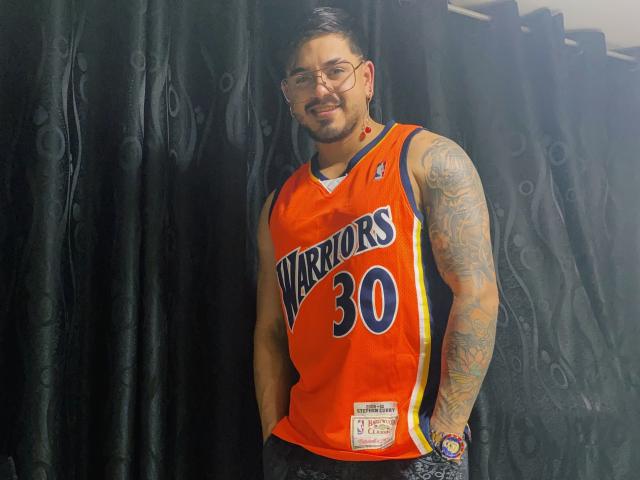 VisiI heeft tatoeages op beide armen, draagt een bril, een oranje Warriors-shirt met het nummer "30" en een rode oorbellen. Hij staat voor zwarte gordijnen en glimlacht..