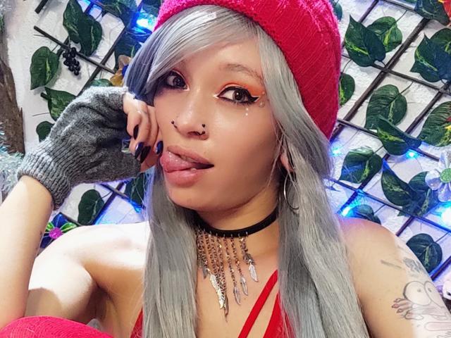 GoddessLufe avec des cheveux argentés, un bonnet rouge, et un gant gris, la langue tirée, un anneau au nez, et un collier en chaîne, montrant des tatouages et un haut rouge, avec des lumières bleues en arrière-plan..