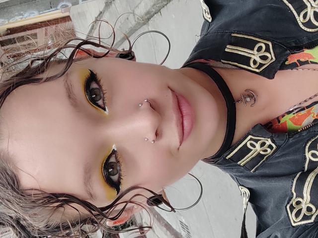 GoddessLufe has yellow eyeshadow, curly wet brown hair, nose piercings, black choker, hoop earrings, and a black jacket with gold details. Her tits are slightly visible.