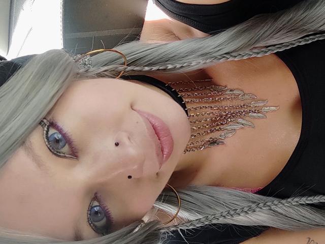 GoddessLufe tiene el cabello trenzado de color gris, ojos azules, un collar negro con plumas de plata y un piercing en la nariz. Lleva una blusa negra y está mirando a la cámara con una sonrisa..