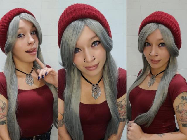 GoddessLufe con i capelli grigi, un berretto rosso, un anello al naso e braccia tatuate, indossa una maglietta rossa, una collana e sorride in tre pose diverse. Il seno è piccolo. La vulva non è visibile..