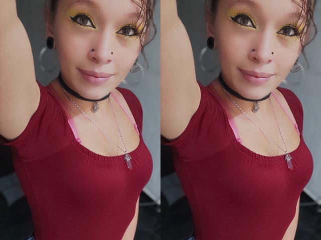 GoddessLufe con ombretto giallo, collana nera, maglietta rossa, reggiseno rosa, seno piccolo, braccio alzato, sorridente, auricolari neri, piercing al naso, capelli castani corti, selfie.