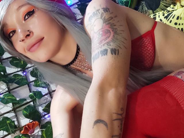 GoddessLufe, avec ses longs cheveux argentés, son soutien-gorge en mesh rouge, ses tatouages, son anneau de nez, son collier et ses bas rouges montants jusqu'à la cuisse, sourit à l'appareil photo, montrant ses seins et ses tatouages sur les bras..