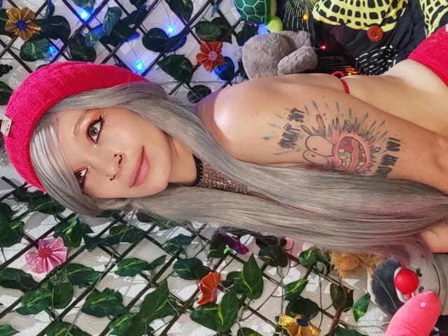 GoddessLufe, avec ses longs cheveux gris, son chapeau rose, son anneau de nez et son tatouage sur le bras droit, nue, montrant ses seins, un sourire mignon, entourée de plantes et de lumières colorées..