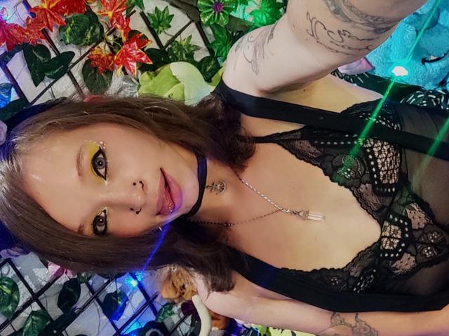 GoddessLufe with wavy brown hair, yellow eyeshadow, black lace lingerie, tattoos, and a choker, sticking out her tongue, showing small tits and a cute face.