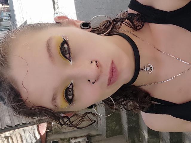 GoddessLufe with yellow eyeshadow, black choker, nose piercings, lip piercing, hoop earrings, and a black top, showing a cute face and curly brown hair.