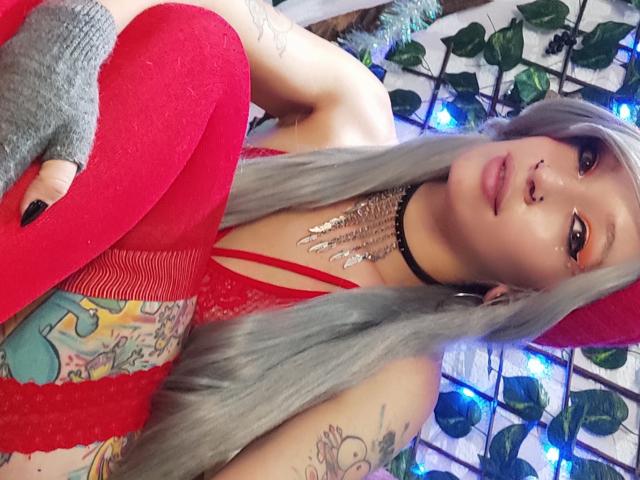 GoddessLufe con cabello largo y gris, lencería de encaje roja y guantes grises sin dedos, mostrando sus bragas rojas, tatuajes y aro en la nariz, llevando un collar negro con una cadena de plata..