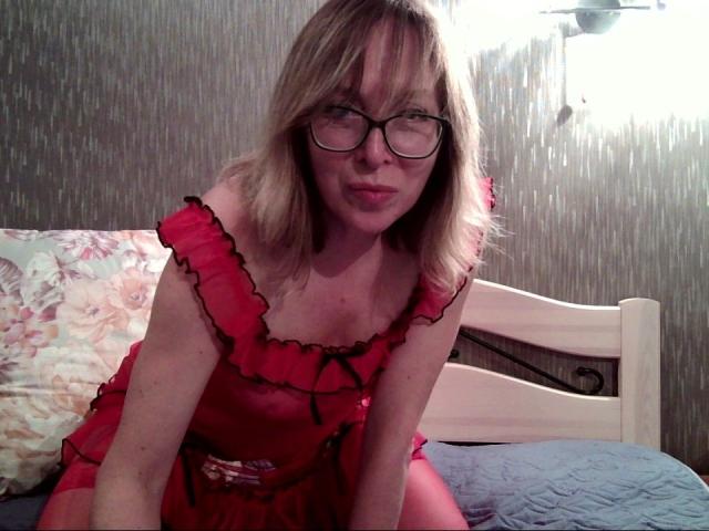 IrunchaArt with blonde hair, glasses, and pink frilled lingerie, leaning forward on a bed, showing her small tits and pouty lips, in a dimly lit room.