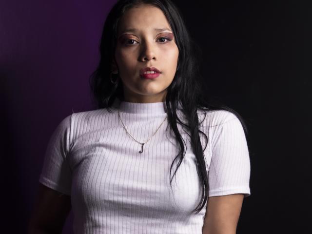 KarollManson has long black hair, wears a white ribbed shirt, and has a small necklace with a black pendant. Her pinkish-red eyes are slightly downcast.