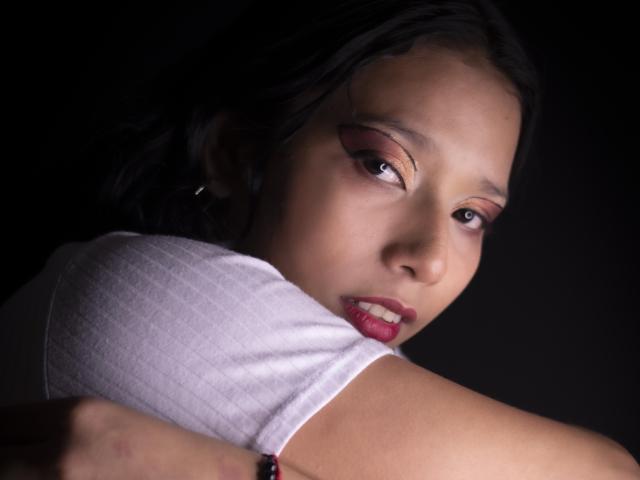 KarollManson with dark hair, brown eyes, and red lipstick, wearing a white shirt, arms crossed, looking sideways with a slight frown, against a dark background.