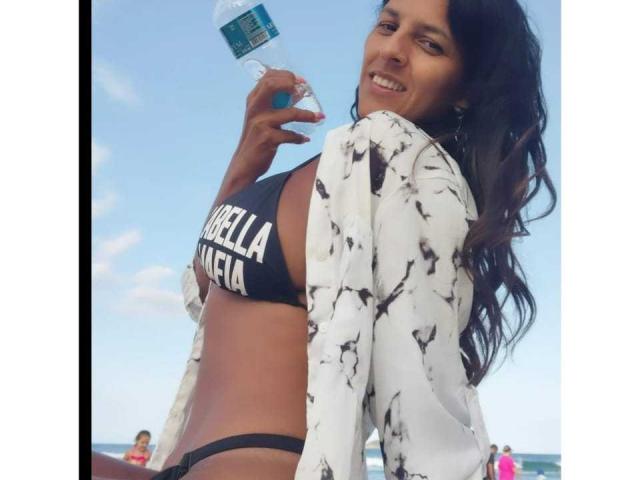 Mulher de pele escura, com longos cabelos pretos ondulados, sorrindo, vestindo um top de biquíni preto, uma sunga preta e uma camisa branca estampada, segurando uma garrafa de água, mostrando os seios..
