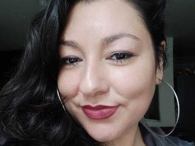 SakotoDika ha i capelli scuri e ondulati, la pelle chiara, l'eyeliner nero, il rossetto rosso e grandi orecchini a cerchio argentati. Sorride leggermente, con un'espressione rilassata e sicura di sé..