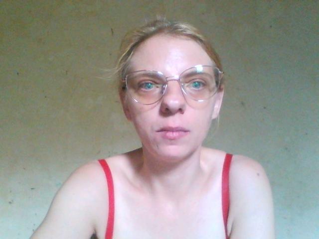 BlondyMarianne tiene la piel pálida, ojos azules y usa gafas transparentes con correas rojas sujetas a sus pechos. Su cabello rubio está despeinado y está mirando directamente a la cámara..