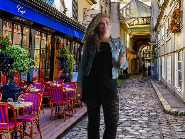 DalilaSuperHot with long brown hair, blue denim jacket, black dress, and black patterned pants, standing on a cobblestone street with colorful chairs and a blue awning.