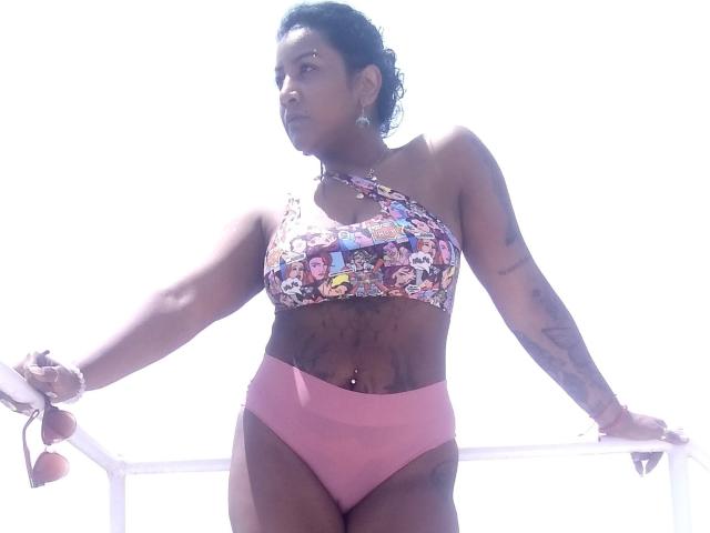 Nahirr, met donkere huid, krullend zwart haar, gekleed in een kleurrijke haltertop en roze hoge bikini broek, staat met haar armen op een witte reling, en houdt roze zonnebrillen vast..