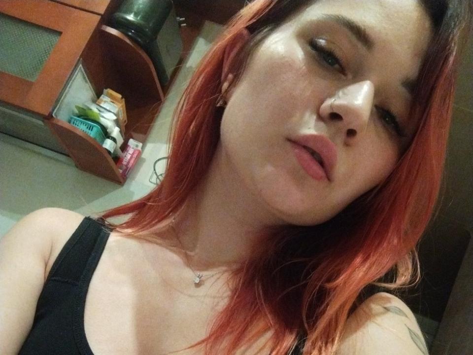 LuckyFanny, con i capelli tinti di rosso, una canottiera nera e una collana, ha un'espressione accigliata e dei liquidi vaginali visibili sul mento, appare leggermente infastidita..