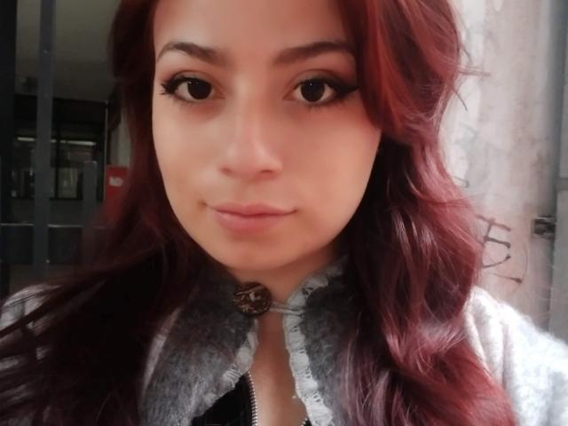 MikuSmile, con cabello rojo, lleva una camisa con cordones, tiene grandes ojos marrones y una sonrisa linda, mostrando un ligero puchero. Sus pechos son pequeños y está mirando a la cámara..