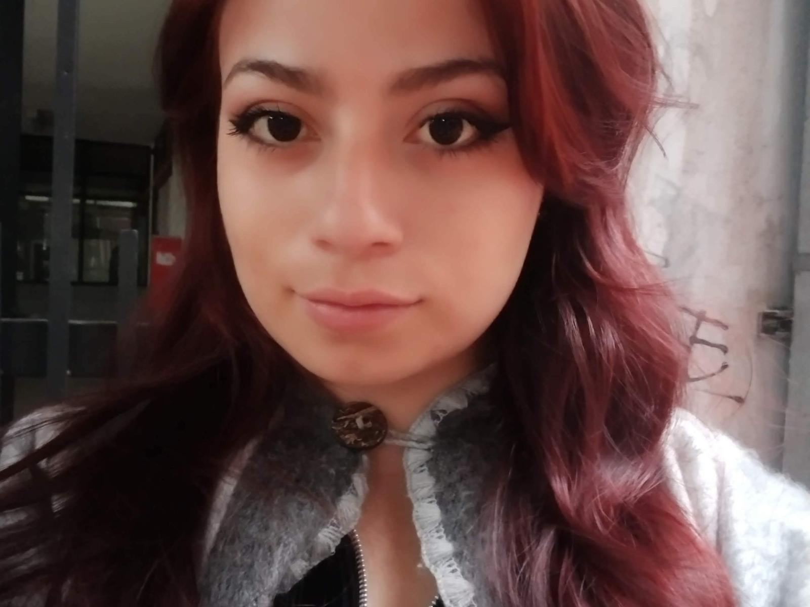 MikuSmile, com cabelo ruivo, vestindo uma camisa com cadarços, tem grandes olhos castanhos e um sorriso fofo, mostrando um leve semblante de descontentamento. Seus seios são pequenos, e ela está olhando para a câmera..