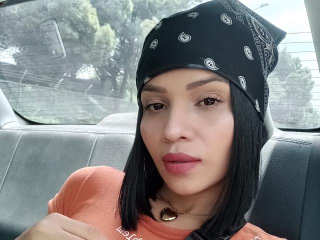 Lily Clark tem a pele marrom média, cabelo preto liso em corte bob, usa um lenço preto com padrões brancos, uma camiseta laranja, brincos de argola e um colar com um coração, e está sentada em um carro..