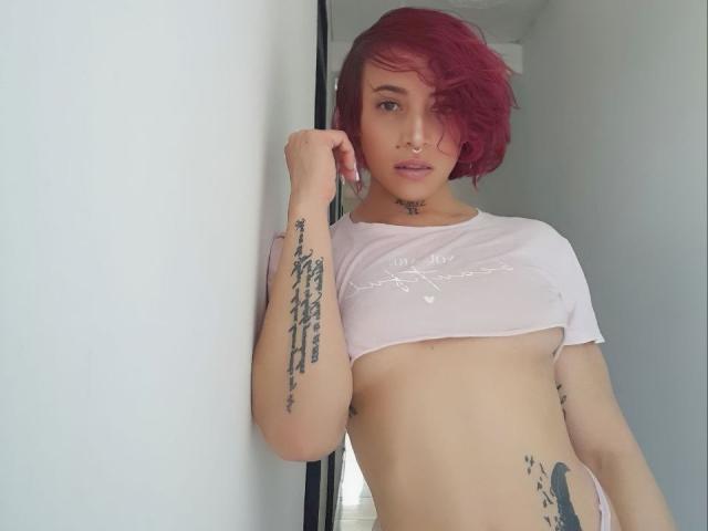 YeiSela met roze haar, een neusring en tatoeages, draagt een witte crop top, waardoor haar borsten zichtbaar zijn, en staat tegen een witte muur, kijkend naar de camera..