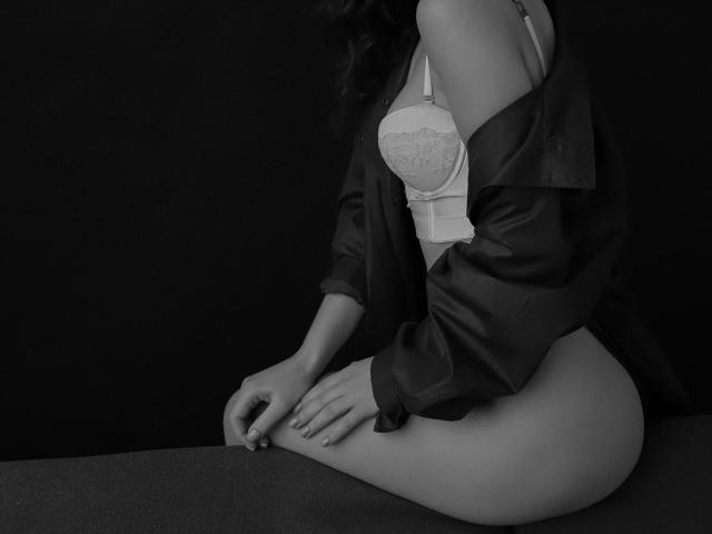 AurikaBriz com camisa de ombro caído, sutiã de renda branco e nádegas expostas, sentada, em preto e branco, mãos nos joelhos, fundo escuro, iluminação suave, elegante, sexy..