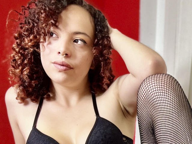 KaterinKlauss met krullend bruin haar, een neusring, een zwarte kanten bh en netpanty, zittend met één arm omhoog, naar de zijkant kijkend, waarbij haar borsten en een opgetrokken knie zichtbaar zijn..