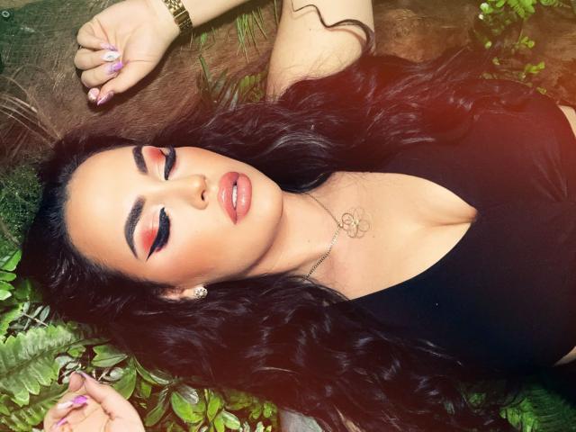 HottyNicole with long black hair, closed eyes, red eyeliner, glossy pink lips, black top, gold bracelet, lying on green foliage, right hand up, left hand down.