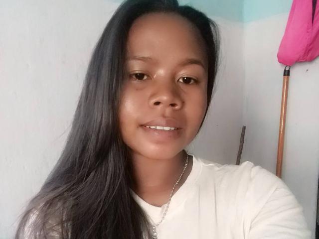 SexyitSlov with long black hair, brown eyes, and a slight smile, wearing a white shirt and silver necklace, has a cute, shiny face and small, straight white teeth.