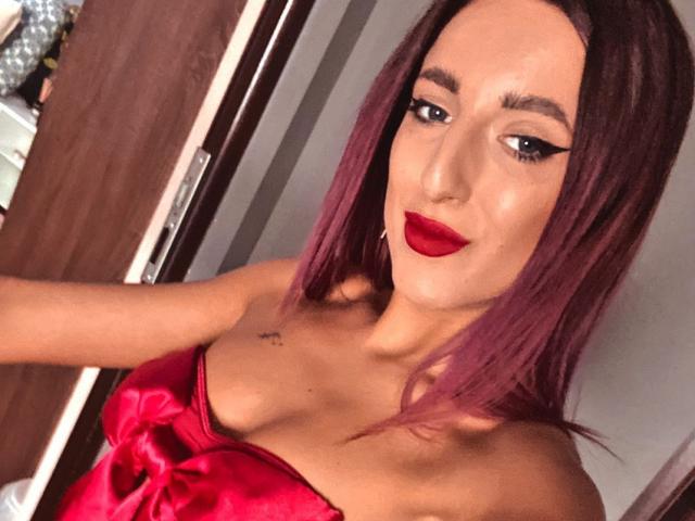 MissAlice69, com batom vermelho, delineador escuro e cabelo roxo, vestindo um vestido de cetim vermelho, mostrando seus pequenos seios e uma tatuagem perto da clavícula..