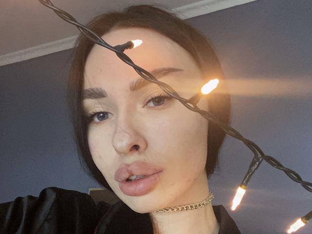 BellaMegan, com cabelo preto curto, usando um colar de corrente dourada, tem luzes de Natal sobre o rosto, lábios rosados levemente entreabertos e olhos azuis, em um quarto com pouca luz..