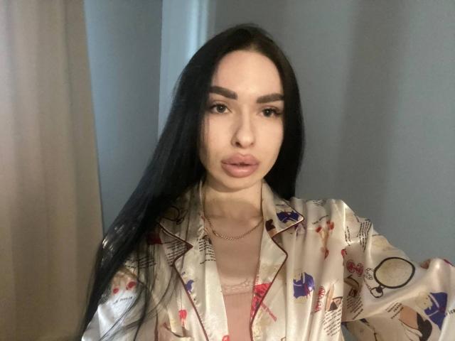 BellaMegan, con su largo cabello negro, vestida con una bata de seda con estampados coloridos, mostrando sus labios rosados y un sujetador de encaje rosa debajo. Lleva un collar delgado..