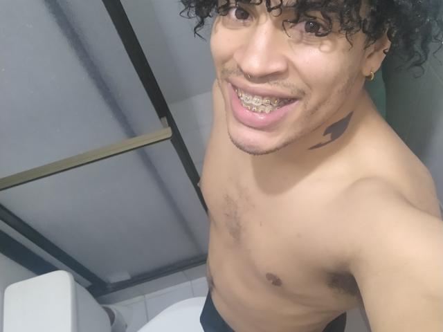 Christopher Wood zonder shirt, met krullend haar, lachend, met metalen tanden zichtbaar, een kleine tatoeage aan de rechterkant, een klein borstgedeelte, zwarte broek, een wit toilet en een glazen douchedeur op de achtergrond..