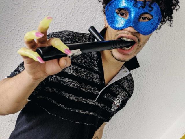 Christopher Wood, con cabello rizado, máscara de lentejuelas azules, camisa a rayas negras, uñas postizas amarillas, sosteniendo un juguete sexual negro en la boca, mirando hacia arriba con la boca abierta..